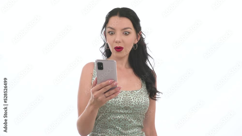 Young shocked, well-groomed woman browsing a mobile phone and emotionally surprised, periodically raising her eyes and looking at the camera. Stunned female bulging her eyes looking into smartphone. Young shocked, well-groomed woman browsing a mobile phone and emotionally surprised, periodically raising her eyes and looking at the camera. Stunned female bulging her eyes looking into smartphone.