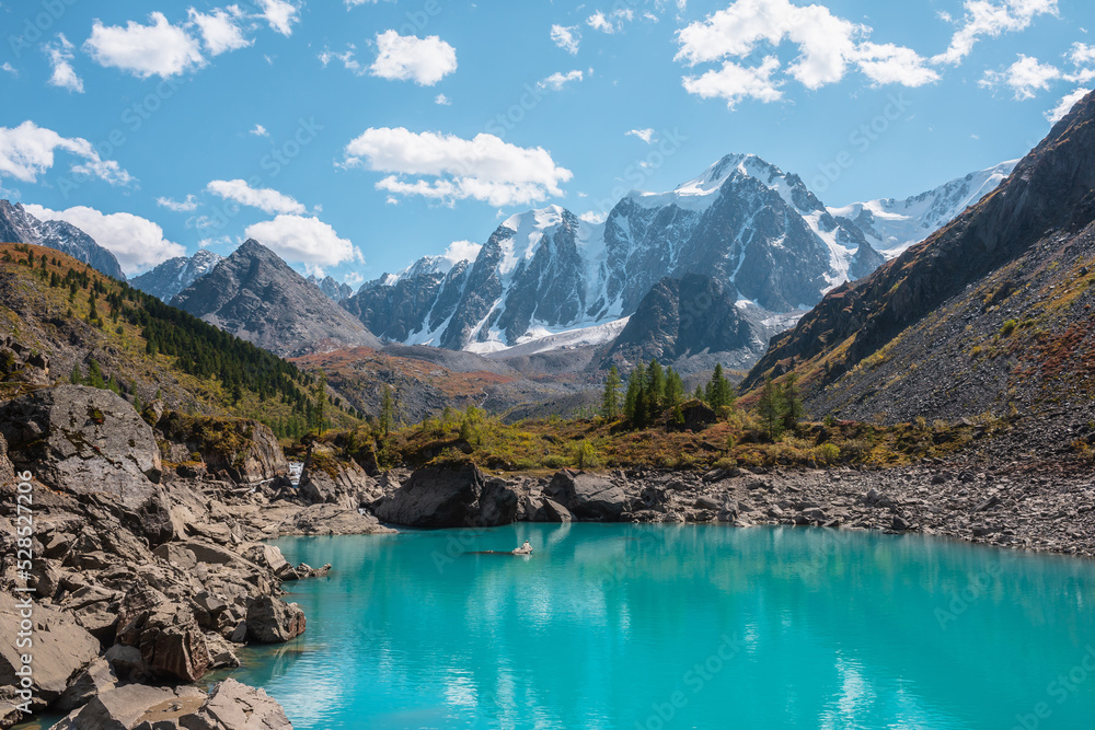 Pure turquoise alpine lake with view to forest hill and giant snow mountains in autumn sunny day. Glacial lake against huge snow covered mountain range in bright sun. Vivid colors in high mountains.