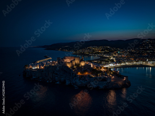 Fotografía aérea nocturna de la ciudad amurallada de Peñíscola iluminada por luces artificiales.