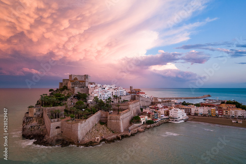 Fotografía aérea de la ciudad amurallada de Peñíscola al atardecer con nubes de colores en el fondo.