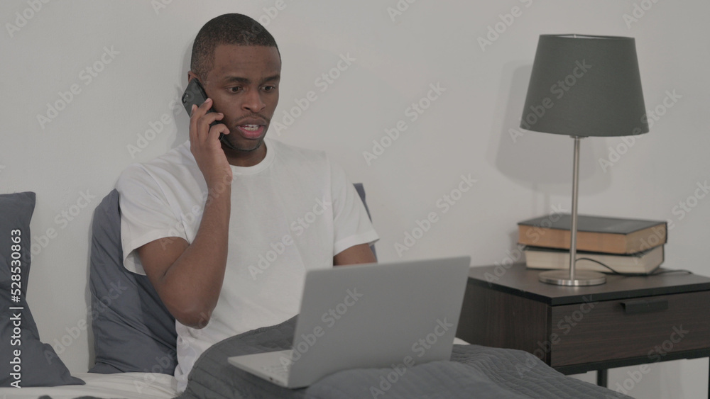 African Man with Laptop Talking on Smartphone, Sitting in Bed African Man with Laptop Talking on Smartphone, Sitting in Bed