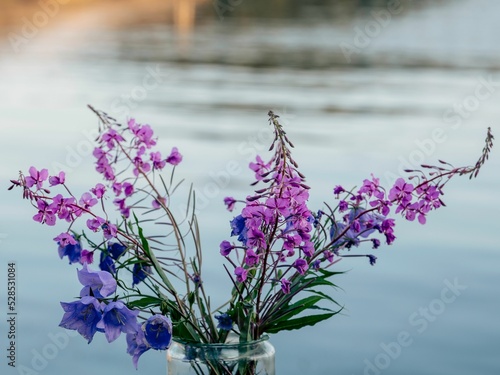 Closeup of fireweed and harebell placed in a glass vase in front of a lake
