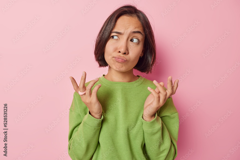 Clueless puzzled Asian woman shrugs shoulders feels hesitant about something spreads hands sideways feels unaware dressed in casual green pullover isolated over pink background. So what to do Clueless puzzled Asian woman shrugs shoulders feels hesitant about something spreads hands sideways feels unaware dressed in casual green pullover isolated over pink background. So what to do