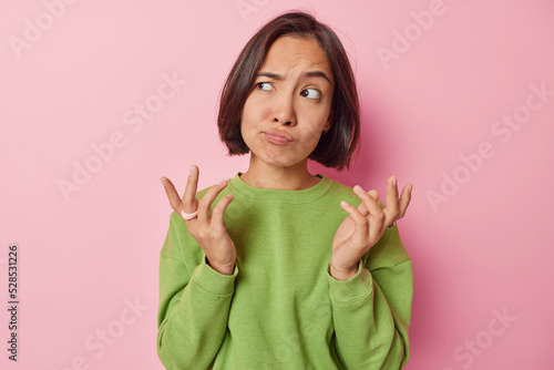 Wallpaper Mural Clueless puzzled Asian woman shrugs shoulders feels hesitant about something spreads hands sideways feels unaware dressed in casual green pullover isolated over pink background