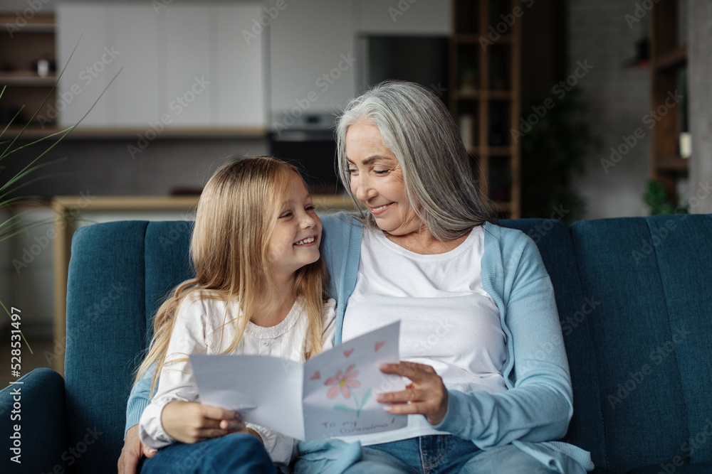 Smiling european small girl gives card to old woman, enjoy moment together in living room interior Smiling european small girl gives card to old woman, enjoy moment together in living room interior