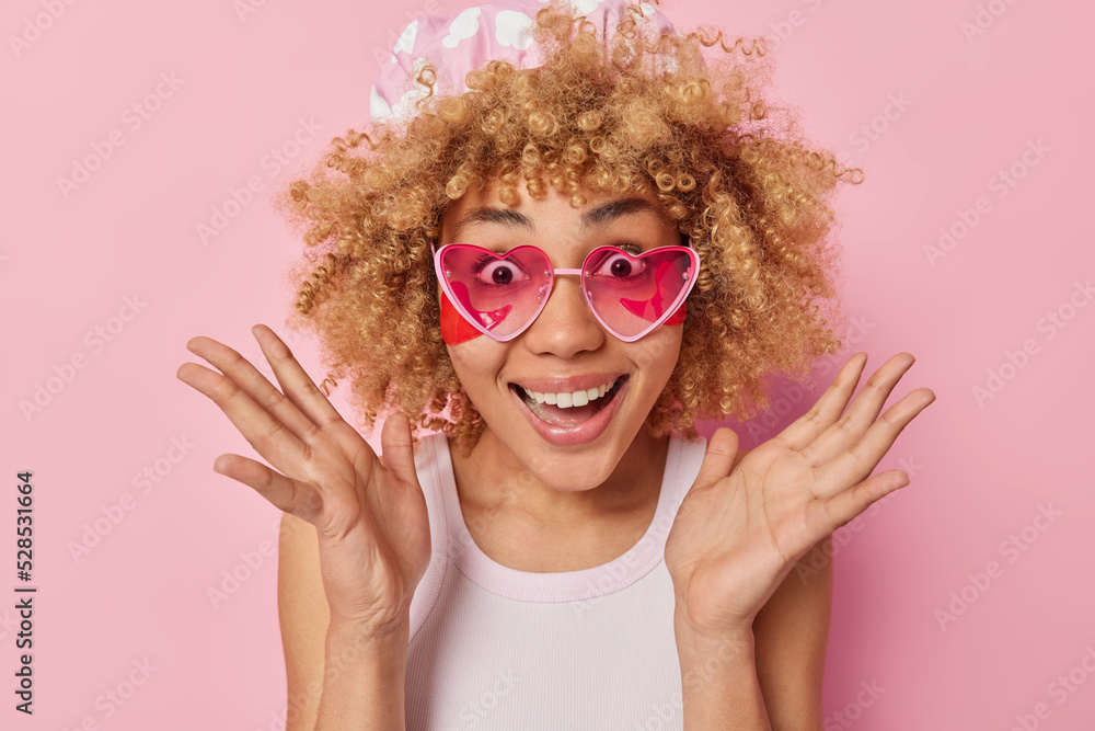 Portrait of good looking young woman spreads palms looks excited smiles broadly applies beauty patches under eyes after taking bath wears shower cap t shirt and pink sunglasses poses indoor. Portrait of good looking young woman spreads palms looks excited smiles broadly applies beauty patches under eyes after taking bath wears shower cap t shirt and pink sunglasses poses indoor.