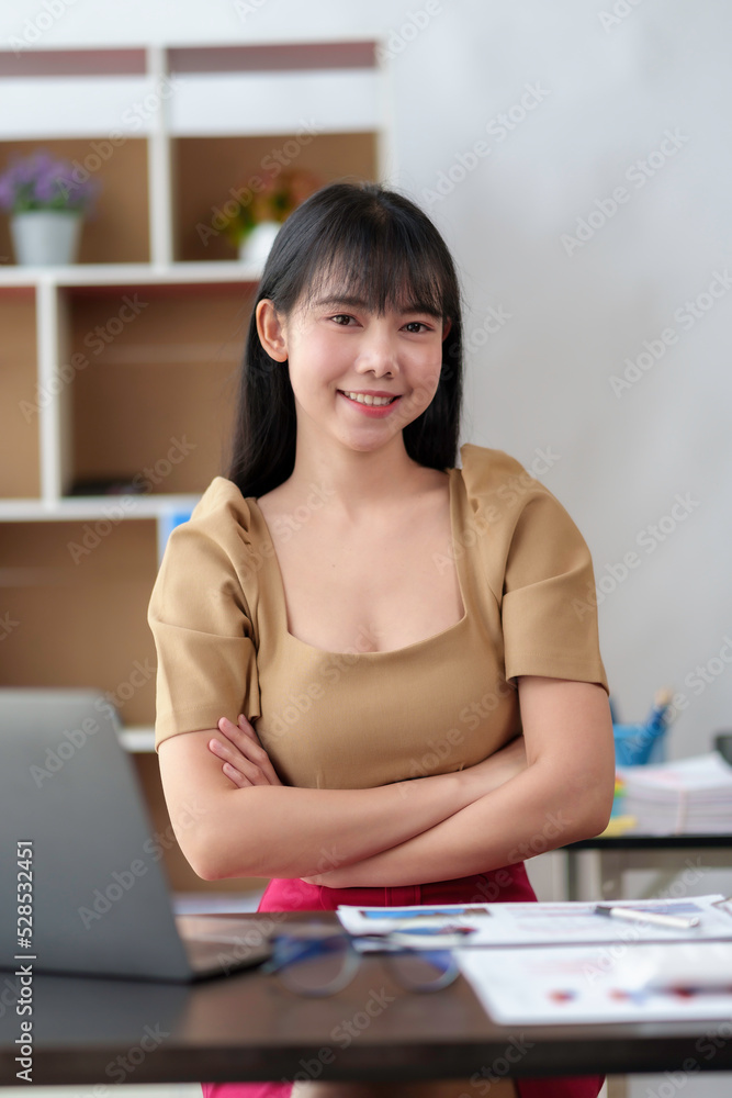 Obraz premium Beautiful Asian businesswoman working on her laptop enjoying work, taking notes, reviewing work and smiling happily looking at the camera.
