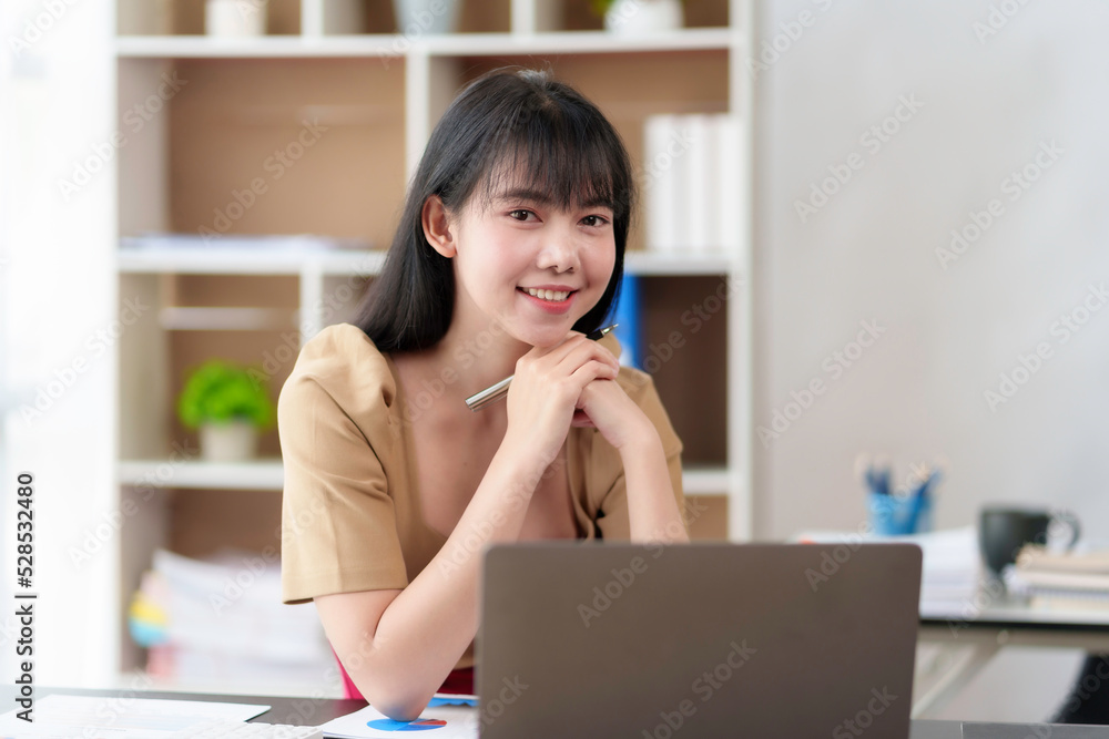 Beautiful Asian businesswoman working on her laptop enjoying work, taking notes, reviewing work and smiling happily looking at the camera. Beautiful Asian businesswoman working on her laptop enjoying work, taking notes, reviewing work and smiling happily looking at the camera.
