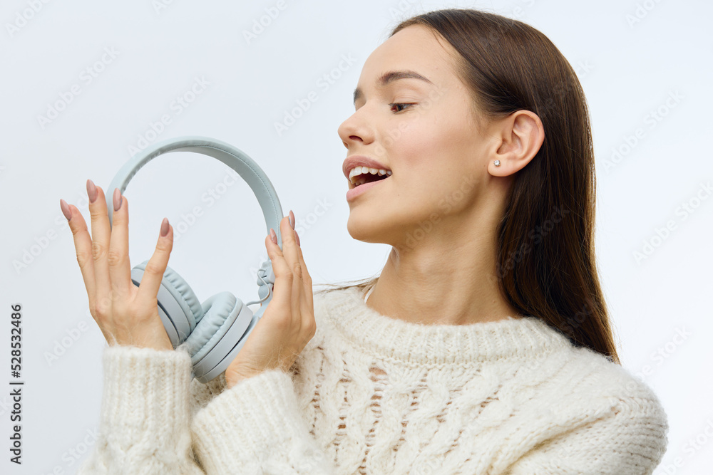 a beautiful, happy woman in a white sweater stands on a gray background holding large headphones in her hands and looking at them with a smile of pleasure