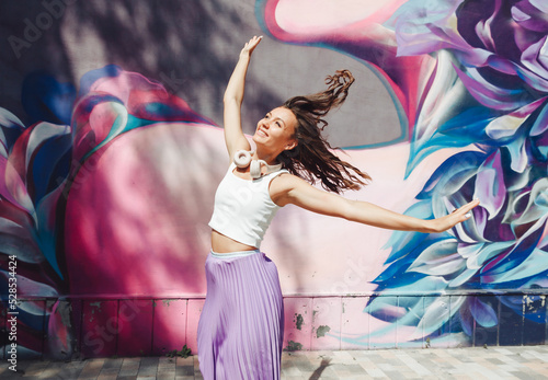 a young excited woman in a top and skirt, wearing headphones, listening to music, walking, dancing with her hands up in the open air against a pink wall.