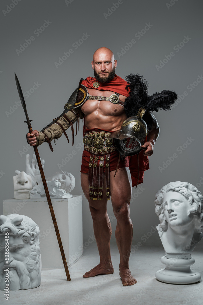 Obraz premium Shot of gladiator dressed in red mantle and armor posing around greek busts.