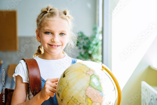 Smiling student girl wearing school backpack and holding globe. Portrait of happy caucasian blond girl inside the primary school. Closeup face of smiling schoolgirl looking at camera.