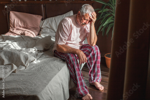 Sad unhappy senior man seated on bed in bedroom