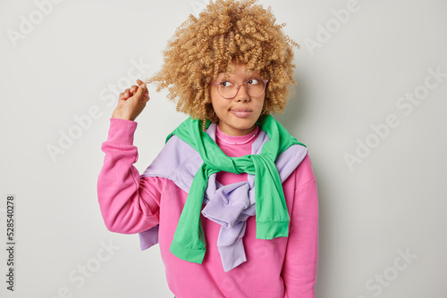 Canvas Print Photo of lovely woman curls hair being deep in thoughts concentrated away considers something wears spectacles casual pink jumper pullovers tied over shoulders isolated over grey background
