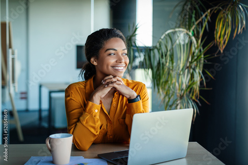 Portrait of dreamy african american businesswoman sitting at desk in office and thinking while working on laptop photo