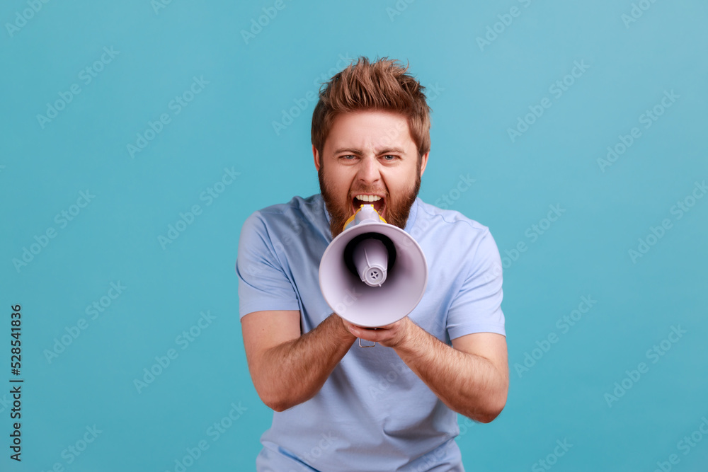 Portrait of bearded man holding megaphone near mouth, loudly speaking, screaming, making announcement, paying attention at social problems. Indoor studio shot isolated on blue background. Portrait of bearded man holding megaphone near mouth, loudly speaking, screaming, making announcement, paying attention at social problems. Indoor studio shot isolated on blue background.
