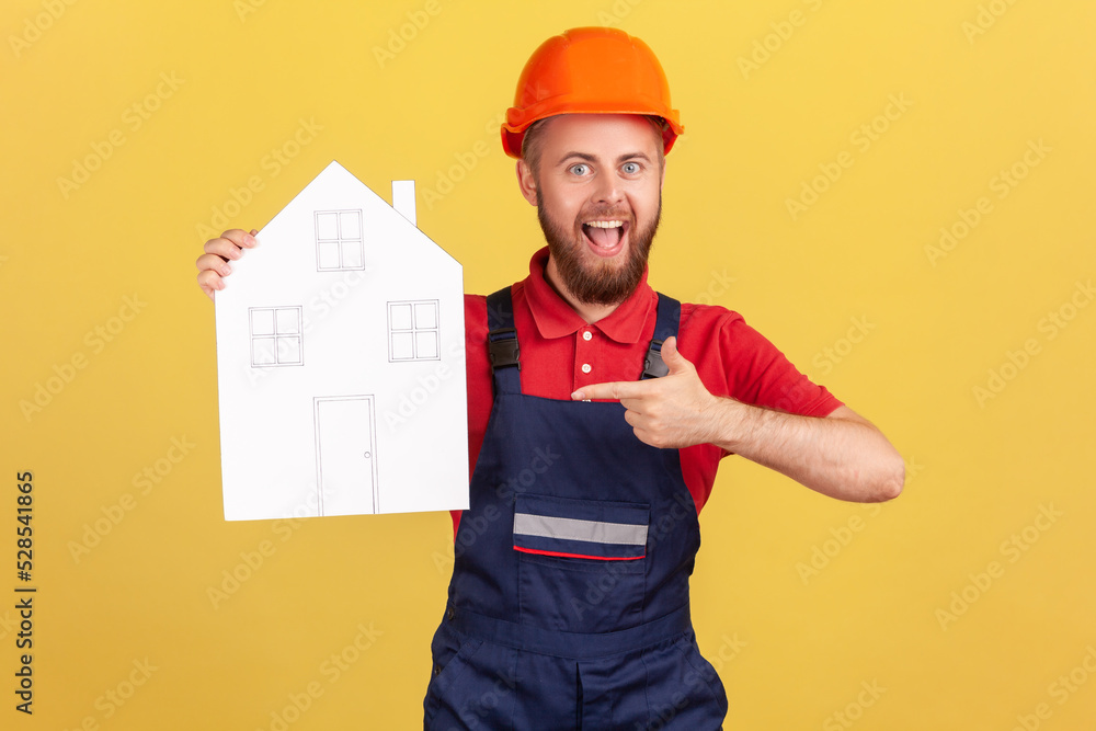 Portrait of builder man pointing at paper house, help in relocation, home repair services, looking at camera with excitement, wearing overalls and cap. Indoor studio shot isolated on yellow background