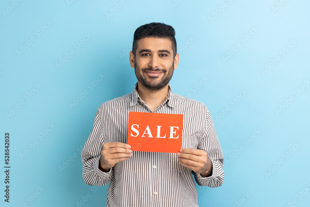 Smiling businessman holding card with Sale word, feeling carefree amused by discounts, Black Friday low prices, wearing striped shirt. Indoor studio shot isolated on blue background. Smiling businessman holding card with Sale word, feeling carefree amused by discounts, Black Friday low prices, wearing striped shirt. Indoor studio shot isolated on blue background.