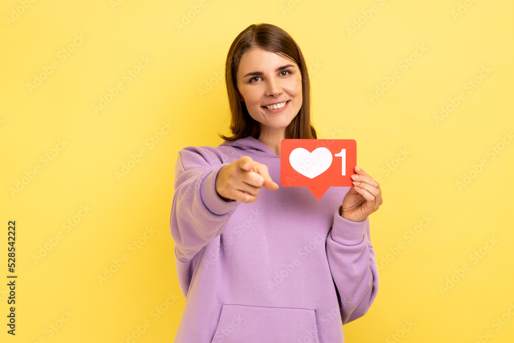 Portrait of dark haired positive woman blogger holding hear like icon and pointing finger at camera with toothy smile, wearing purple hoodie. Indoor studio shot isolated on yellow background. Portrait of dark haired positive woman blogger holding hear like icon and pointing finger at camera with toothy smile, wearing purple hoodie. Indoor studio shot isolated on yellow background.