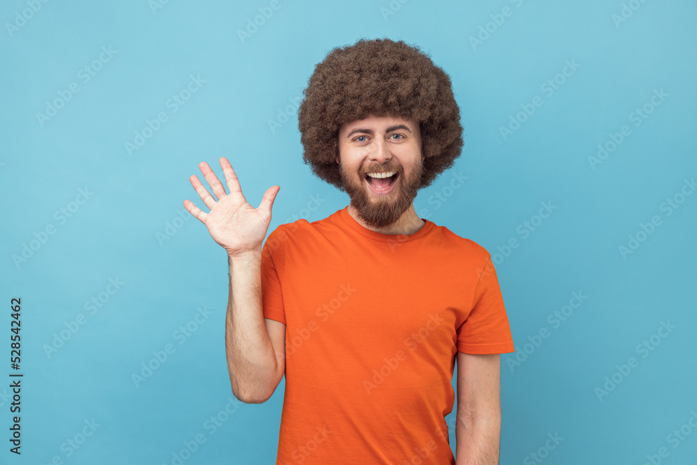 Portrait of cheerful friendly man with Afro hairstyle wearing orange T-shirt saying hi and waving hand, greeting, looking at camera. Indoor studio shot isolated on blue background. - obrazy, fototapety, plakaty 