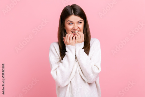Pretty brunette woman biting nails  being nervous terrified  feeling frightened  anxiety disorder  wearing white casual style sweater. Indoor studio shot isolated on pink background.