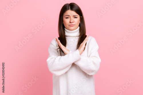 Portrait of determined brunette young woman showing x sign with crossed hands, stop, this is the end, wearing white casual style sweater. Indoor studio shot isolated on pink background.