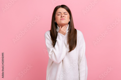 Portrait of sad ill woman touching her neck, suffering sore throat, viral infection or flu symptoms, wearing white casual style sweater. Indoor studio shot isolated on pink background.