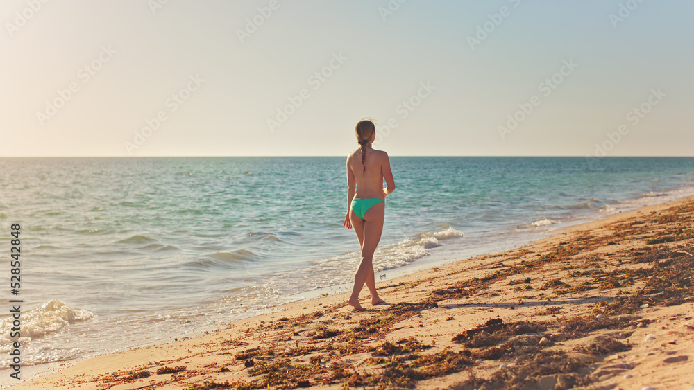 Lean athletic young woman in green blue bikini briefs, walking on sunny beach, view from behind Lean athletic young woman in green blue bikini briefs, walking on sunny beach, view from behind