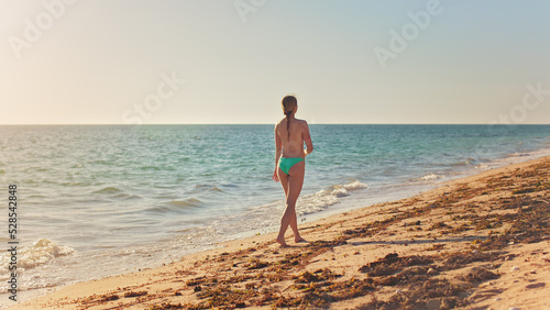 Lean athletic young woman in green blue bikini briefs, walking on sunny beach, view from behind