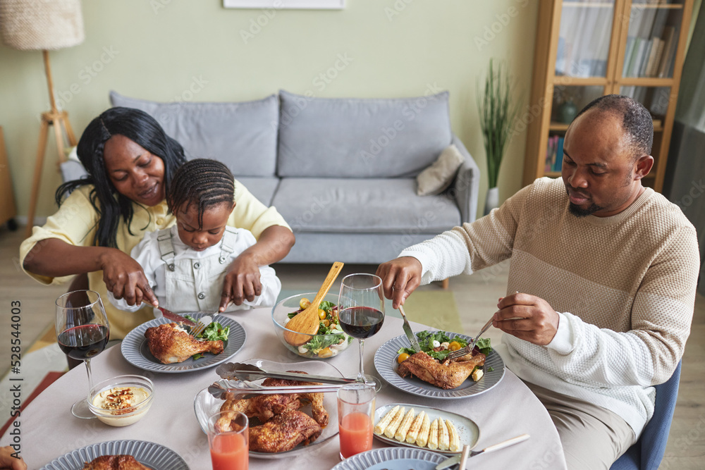 African woman feeding her child with roast chicken during dinner at table in living room African woman feeding her child with roast chicken during dinner at table in living room