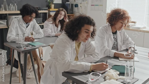 Tracking in of diverse college students sitting at desks in classroom, writing summary on chemistry lecture, professor holding beamer with purple liquid