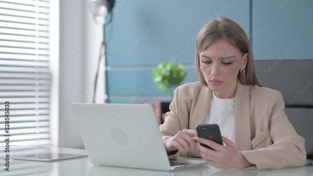 Businesswoman using Smartphone while using Laptop in Office Businesswoman using Smartphone while using Laptop in Office