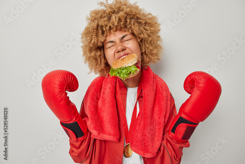 Depressed curly haired female boxer bites delicious sandwich breaks diet and eats fast food aftr workout dressed in sportswear prepares for boxing competition poses indoors against grey background photo