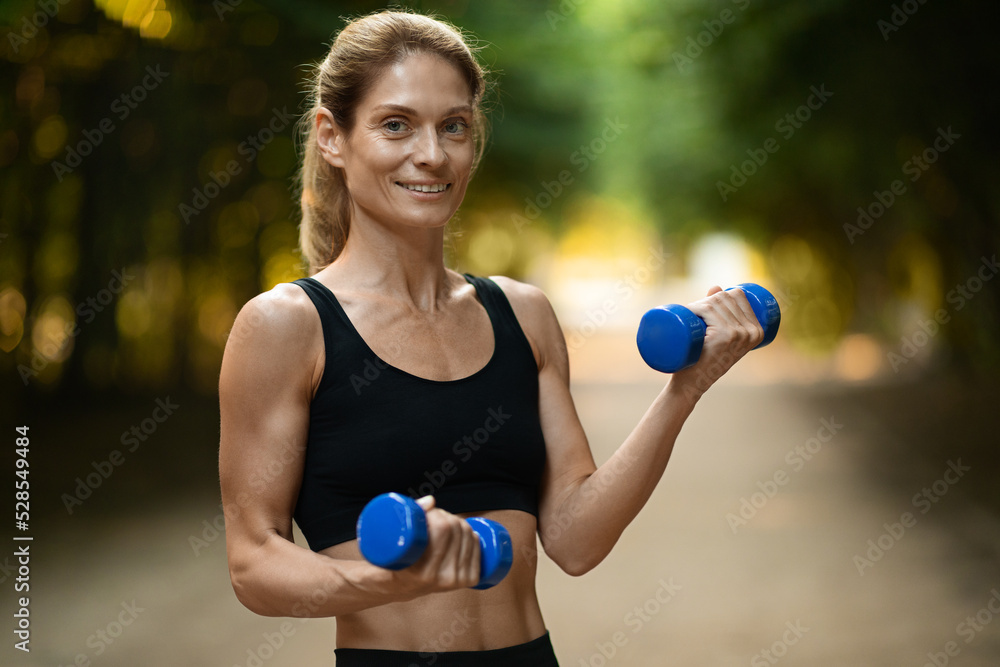 Cheerful motivated sporty woman exercising with dumbbells in park Cheerful motivated sporty woman exercising with dumbbells in park