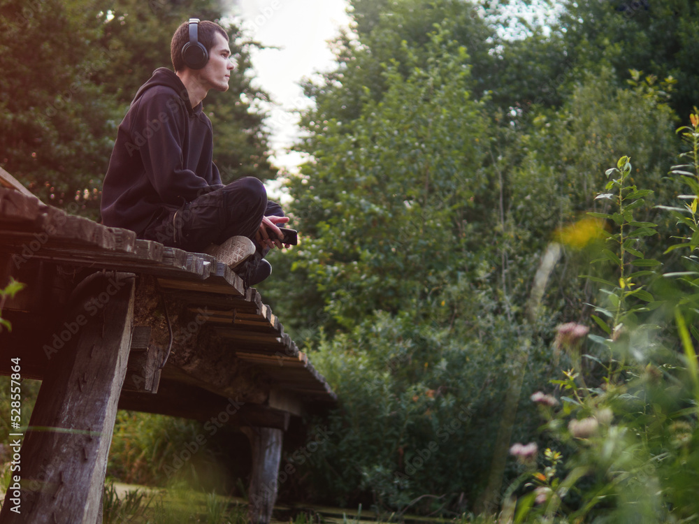 Fototapeta premium A young man is sitting on an old wooden bridge and listening to music with headphones