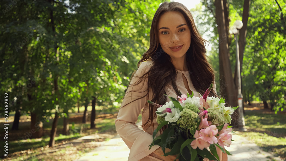Cheerful woman looking at camera while holding bouquet in green park. Cheerful woman looking at camera while holding bouquet in green park.