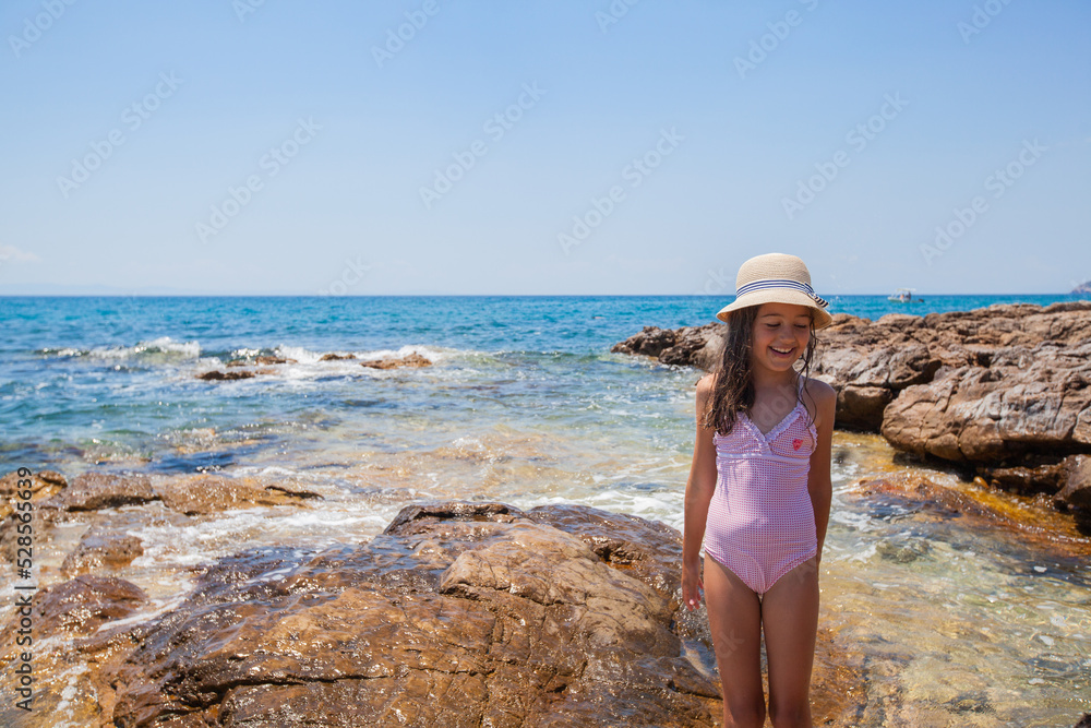 Happy little girl with straw hat at rocks shoreline during summer vacation. Portrait of smiling child at beach. Sunny day. Beautiful seascape in background.