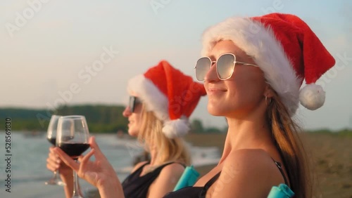 Two young beautiful slender Caucasian women in santa clasus hat, for Christmas, sit on chairs on beach, by sea or ocean and drink wine. They strike glasses rejoicing, celebrating and laughing. photo