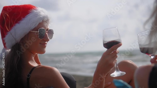 Two young beautiful slender Caucasian women in santa clasus hat, for Christmas, sit on chairs on beach, by sea or ocean and drink wine. They strike glasses rejoicing, celebrating and laughing. photo