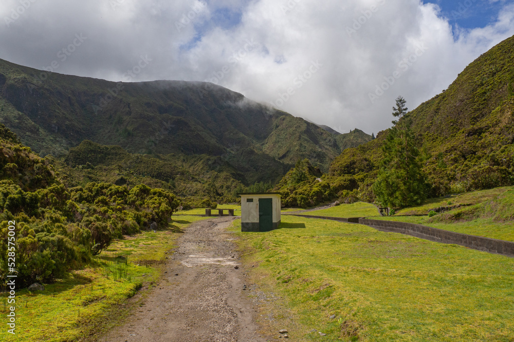Small hidden house isolated with path in green empty field surrounded by nature, trees, hills, mountains and forest during summer in são miguel island, azores, açores, portugal	
