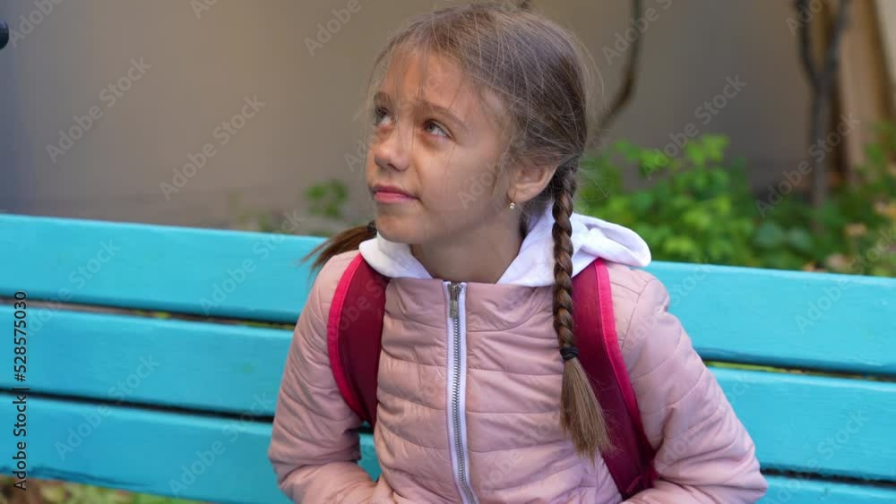 Thoughtful schoolgirl girl in a warm jacket sits on a bench near the school and looks around in anticipation. Thoughtful schoolgirl girl in a warm jacket sits on a bench near the school and looks around in anticipation.