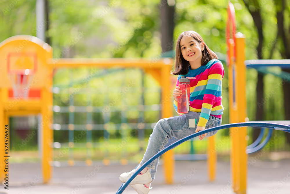 Obraz premium Adorable smiling little girl, 5-6 years old, sitting with a plastic bottle of water on the playground