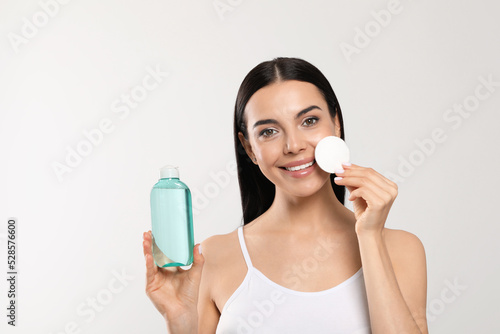 Young woman with micellar water and cotton pad on light grey background