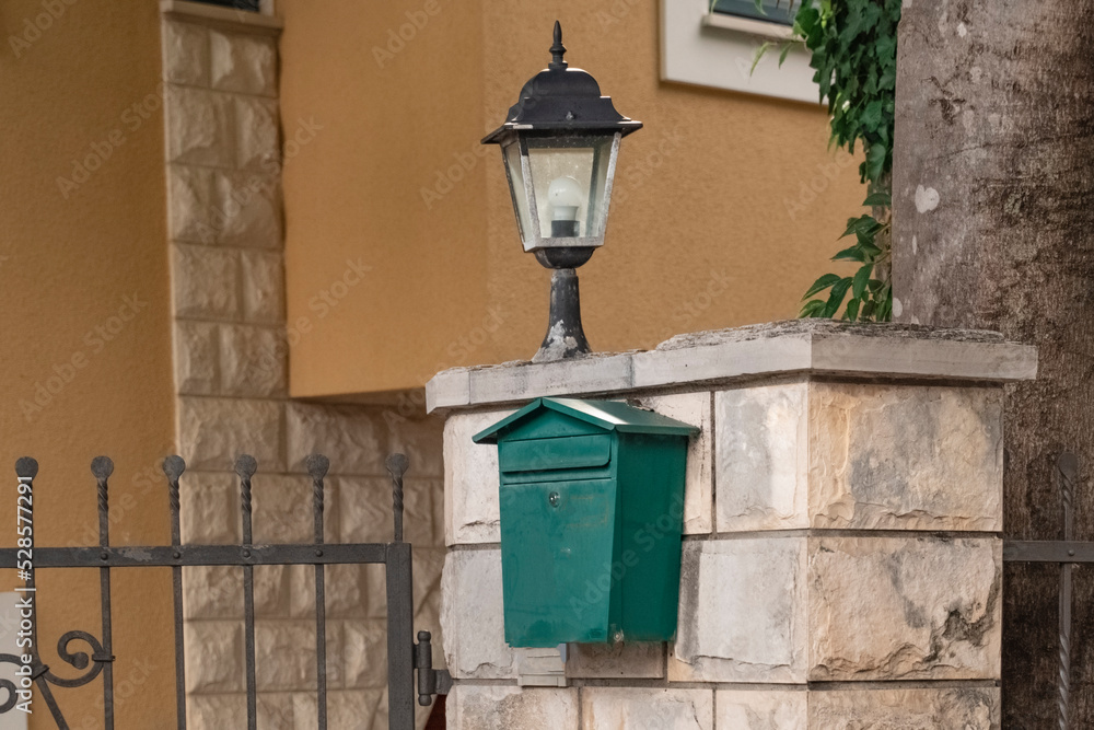 An old green mailbox hangs on a stone fence near an old street lamp. Soviet mailbox. A place to store mail.