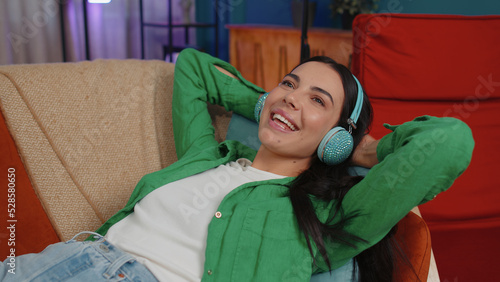 Young woman in wireless headphones relaxing lying on sofa at home choosing listening favorite energetic disco rock n roll dancing music in smartphone. People weekend activities. Happy overjoyed girl