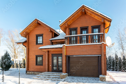 Scenery facade front view of modern wooden luxury timber eco house villa with garage door snowcapped by white fresh snow at cold sunny winter day. Real estate building construction project exterior