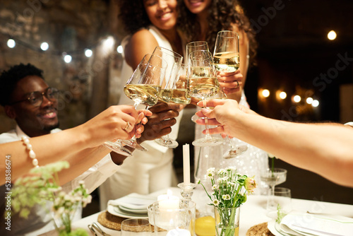Hands of wedding guests and two intercultural brides clinking with flutes of champagne over festive table while enjoying party