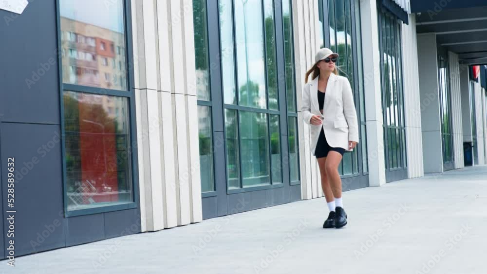 Young fashionable modern lady walking by the street. Model wearing white jacket, black cycling shorts, cap and shoes outdoors. Young fashionable modern lady walking by the street. Model wearing white jacket, black cycling shorts, cap and shoes outdoors.