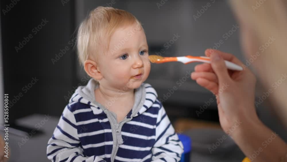 Front view adorable cute little girl eating baby food with unrecognizable woman feeding toddler. Pretty Caucasian child enjoying tasty meal sitting in kitchen at home with mother Front view adorable cute little girl eating baby food with unrecognizable woman feeding toddler. Pretty Caucasian child enjoying tasty meal sitting in kitchen at home with mother