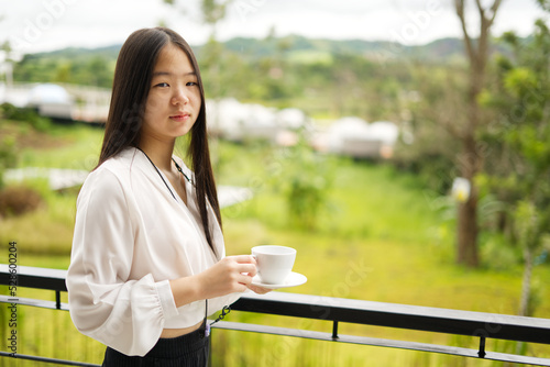 An Asia girl in white shirt is standing on balcony and holding a white cup with blur background and copy space from Thailand.